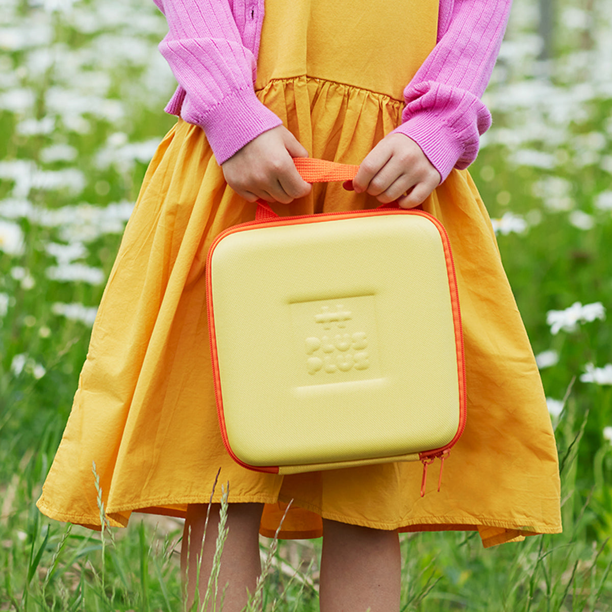 A child in a yellow dress holding the Plus-Plus BIG travel case outdoors in a field.