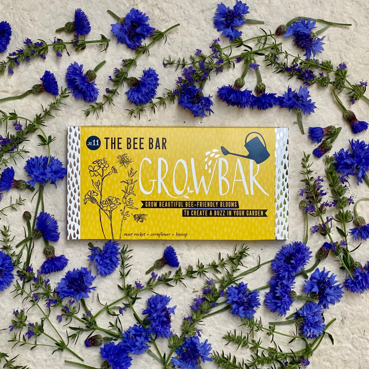A yellow Growbar package labelled "The Bee Bar" surrounded by vibrant blue cornflowers, designed to grow bee-friendly blooms.