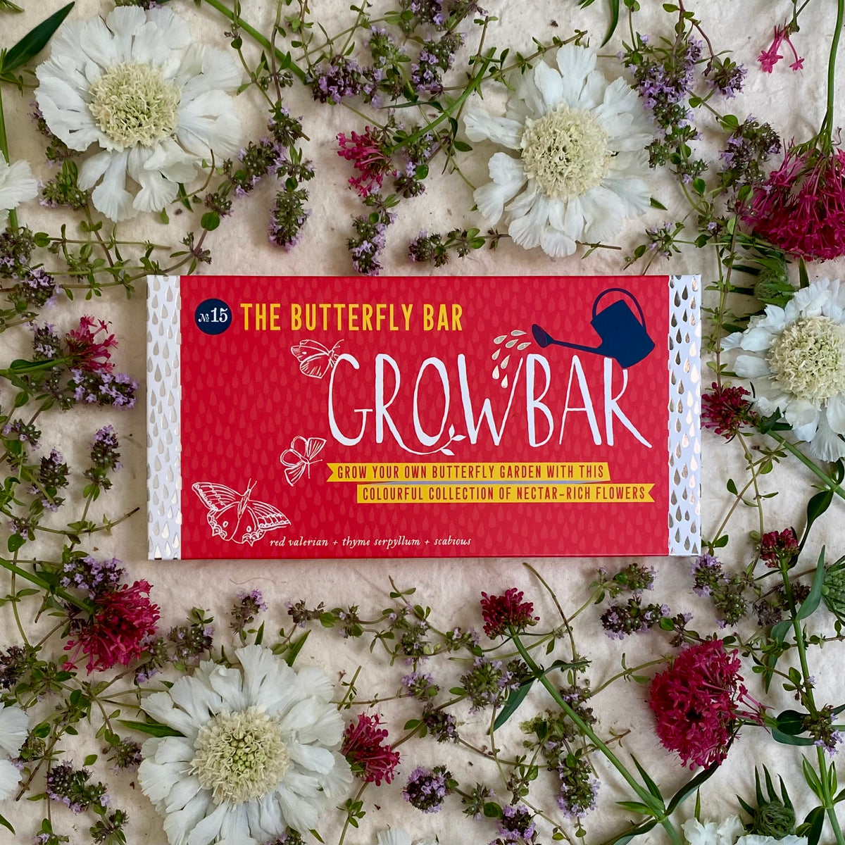 A red Growbar package labelled "The Butterfly Bar" surrounded by delicate white and pink flowers, perfect for growing a butterfly-friendly garden.