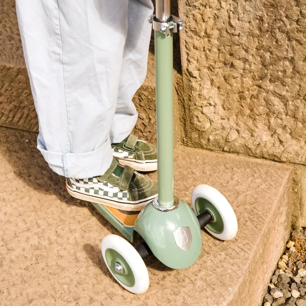 Close-up of a child standing on a sage green 3-wheeled scooter on stone steps.