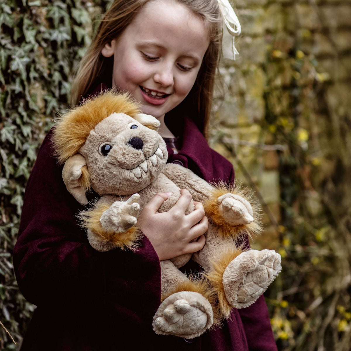 Young girl wearing a maroon coat smiling as she cradles a plush beige creature toy with fluffy yellow hair.