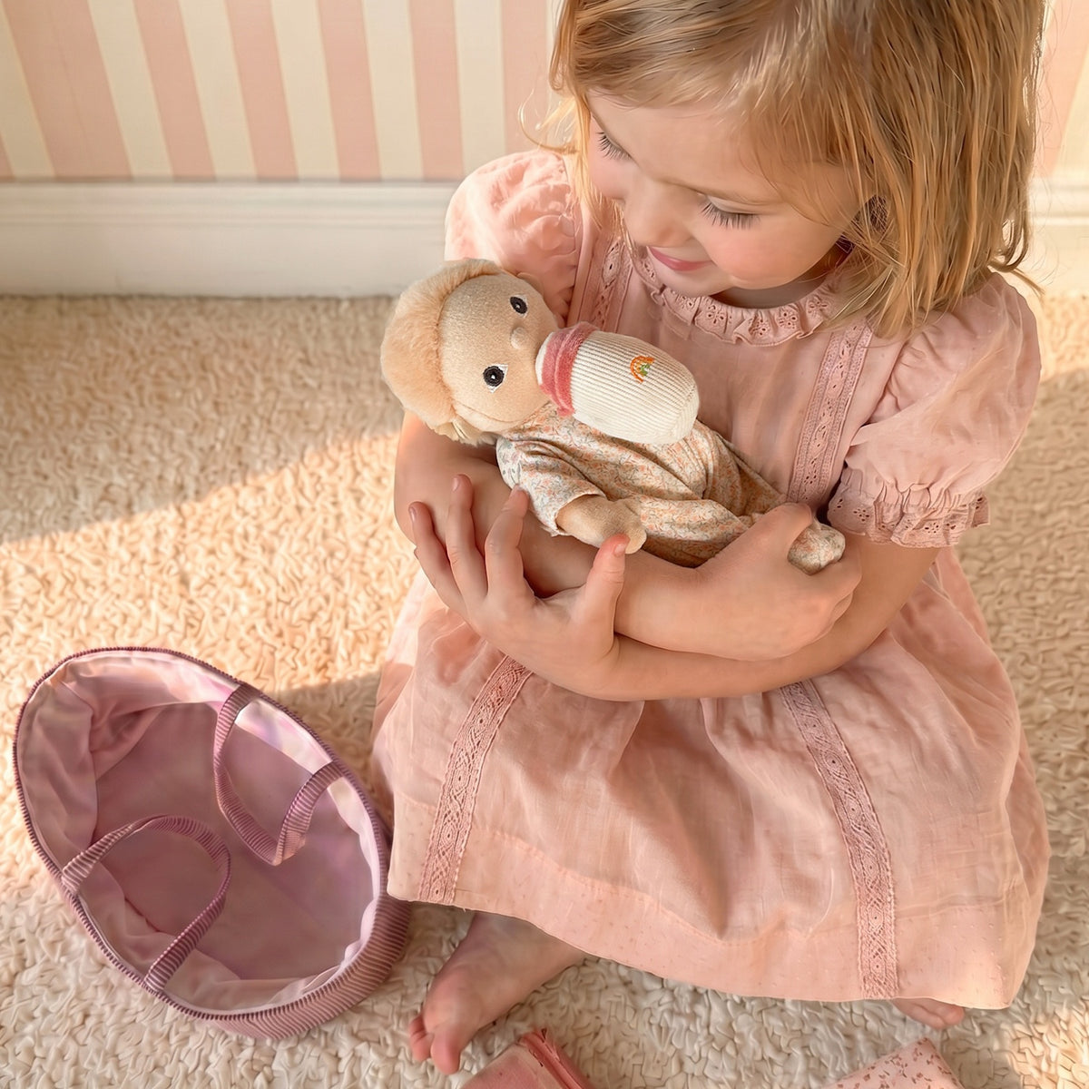 Young girl in a pink dress cradling an Olli Ella Dinky Dinkum Moppet doll with a bottle.