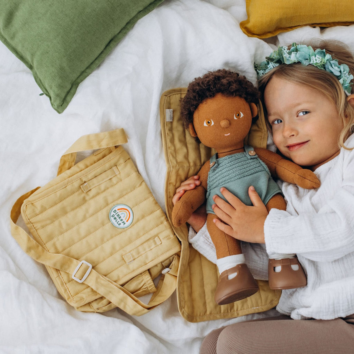 A young girl lying on a bed next to a honey yellow doll changing bag and a Dinkum Doll on a mat.