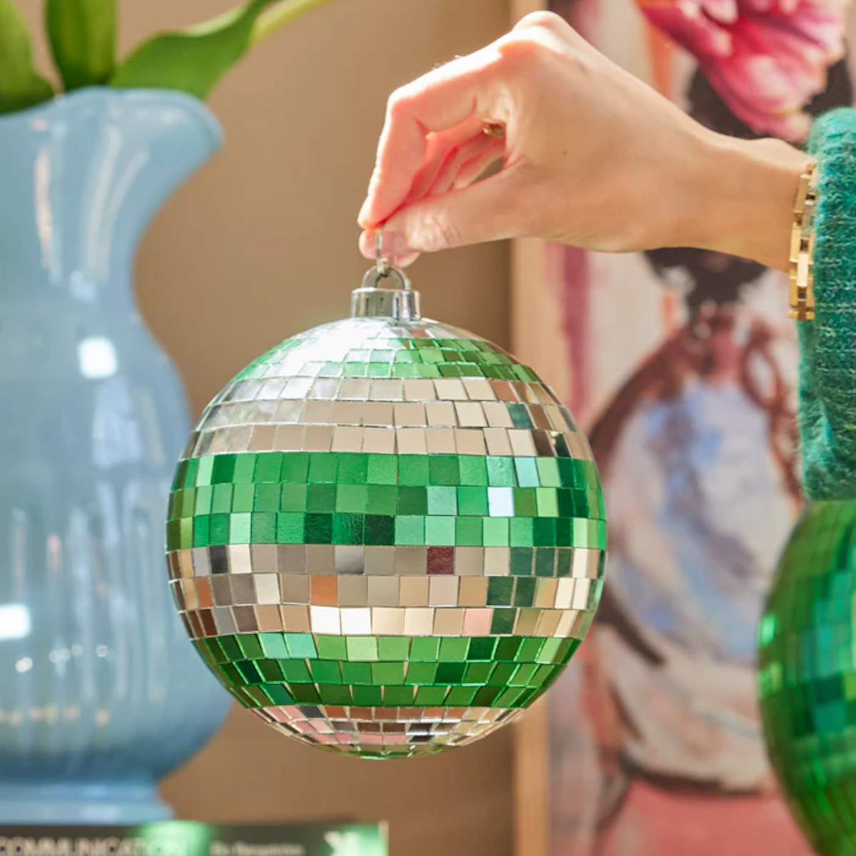A hand holds a green and silver disco ball ornament. Nearby, a blue vase houses green leaves, and art books are stacked on a wooden surface. The mood is festive.