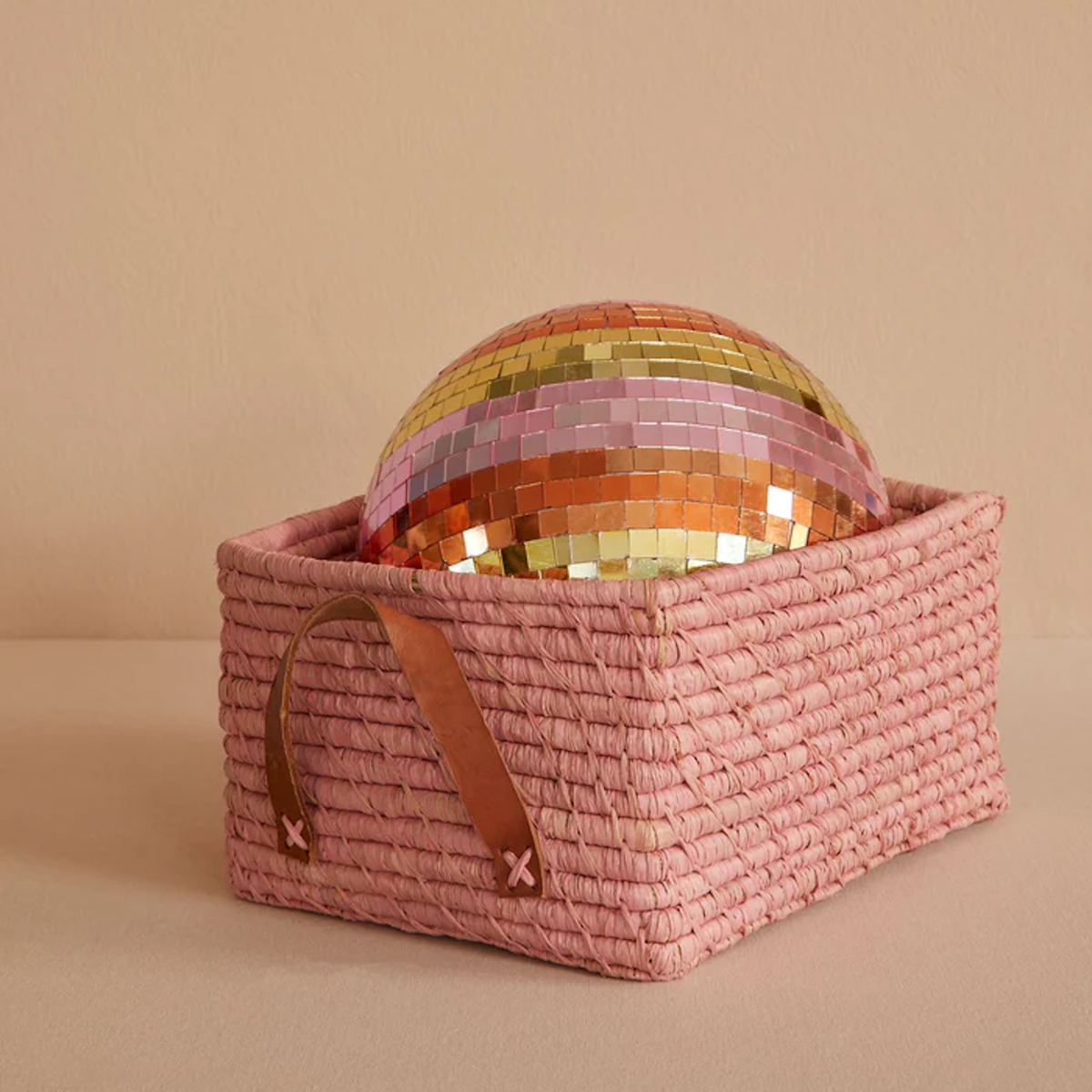 A colorful mirrored disco ball sits in a textured pink woven basket with leather handles against a soft beige background, creating a playful and stylish vibe.