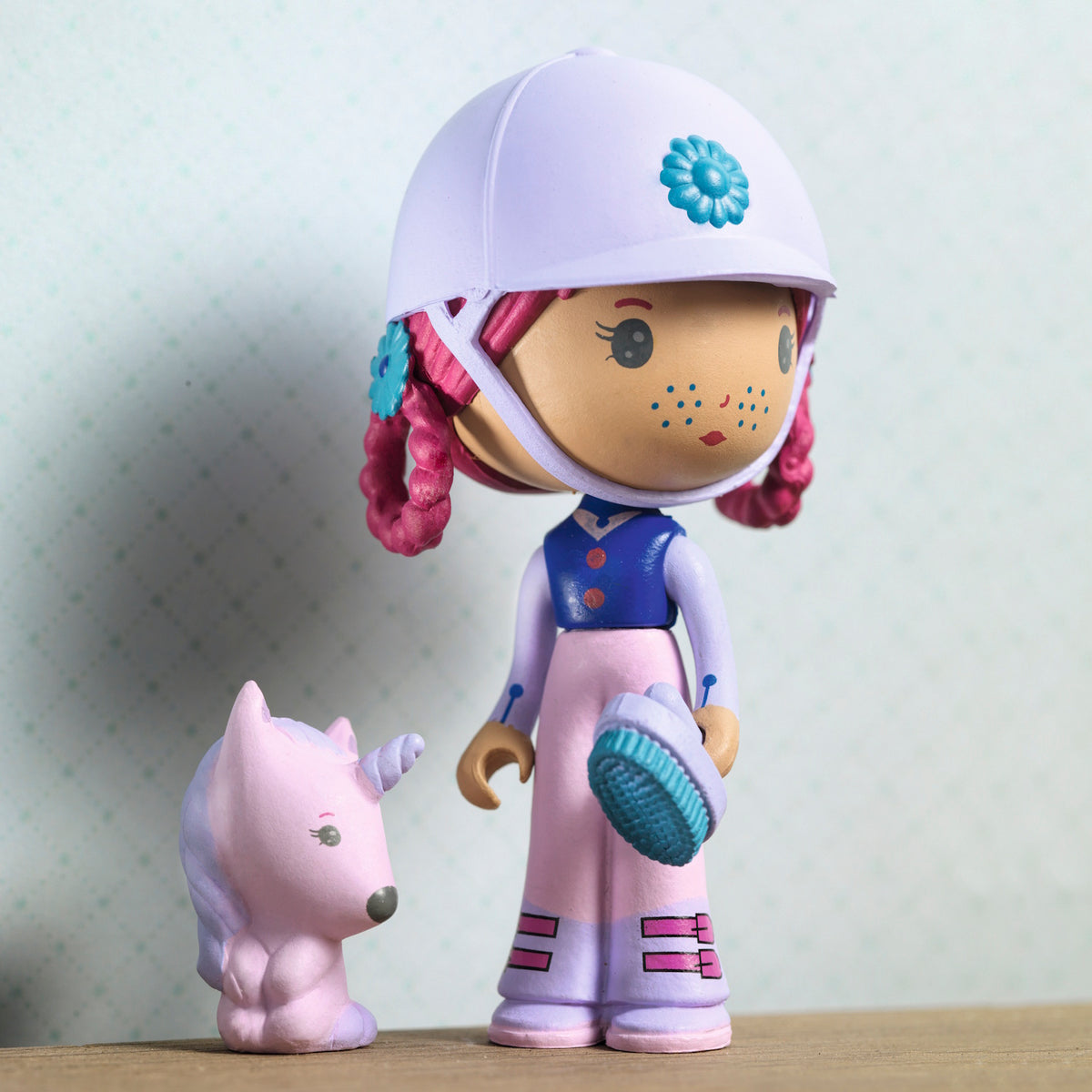 A doll with pink hair and a white helmet, holding a blue brush, stands beside a small purple unicorn. The doll wears a blue and pink outfit.