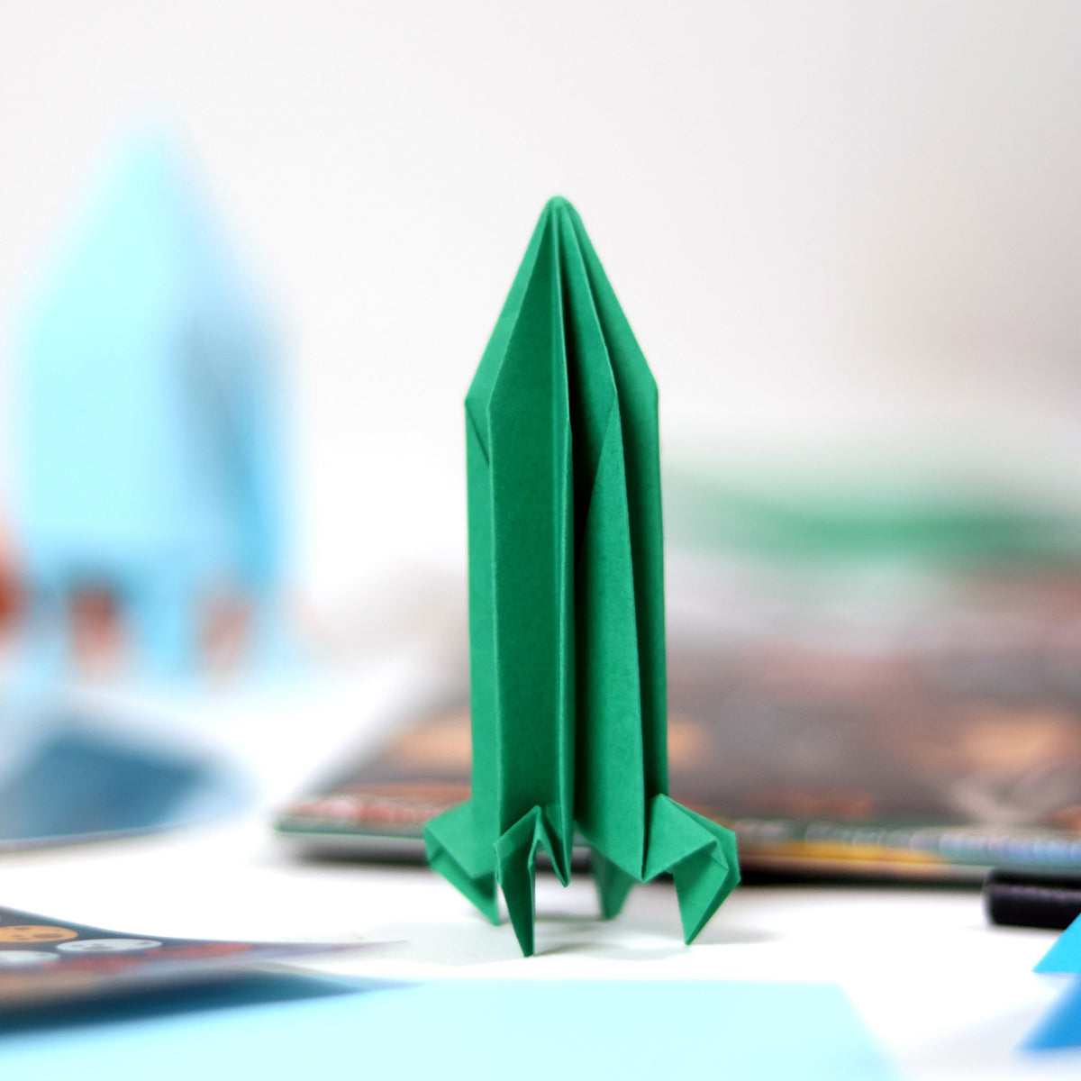 Green origami rocket standing upright on a white surface with craft supplies in the background.