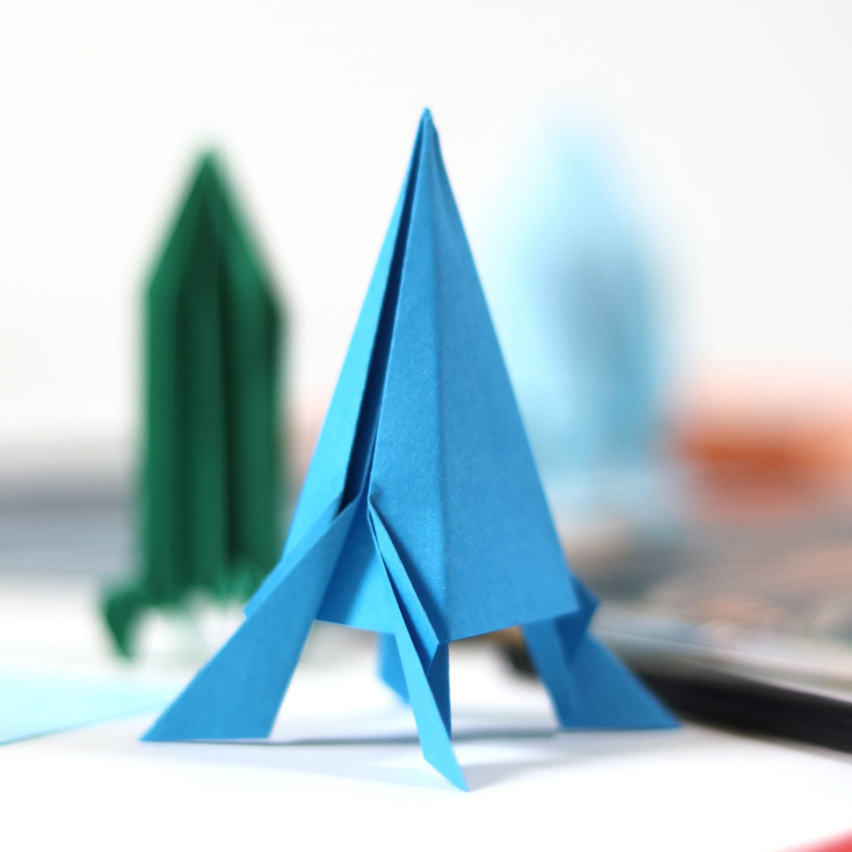 A blue folded origami rocket standing upright with a green paper rocket blurred in the background.