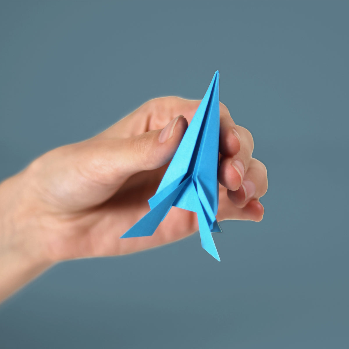 A hand holding a blue folded origami rocket against a solid blue background.
