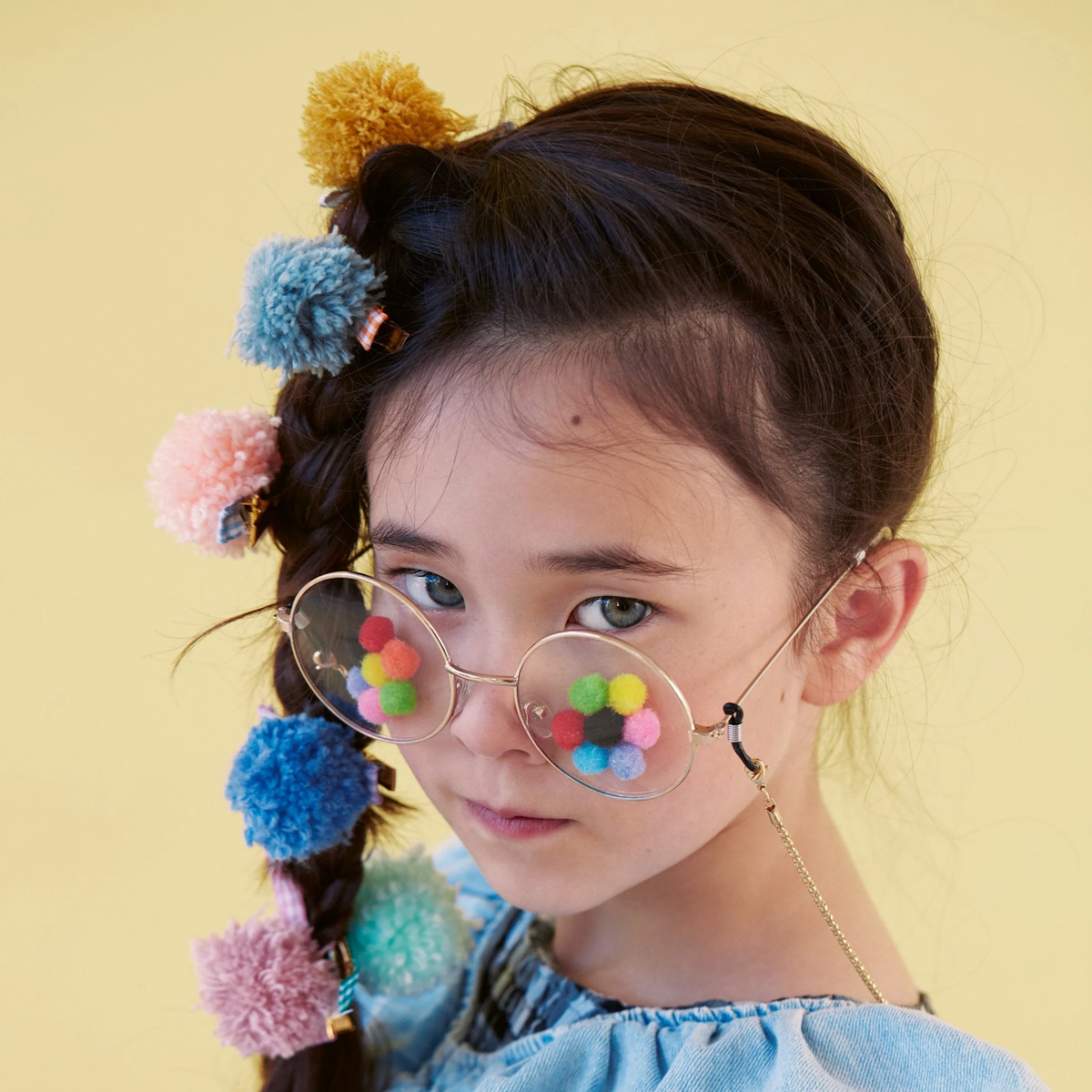 A young girl wearing pom-pom hair clips in her braided hair and matching round glasses decorated with small pom-poms.