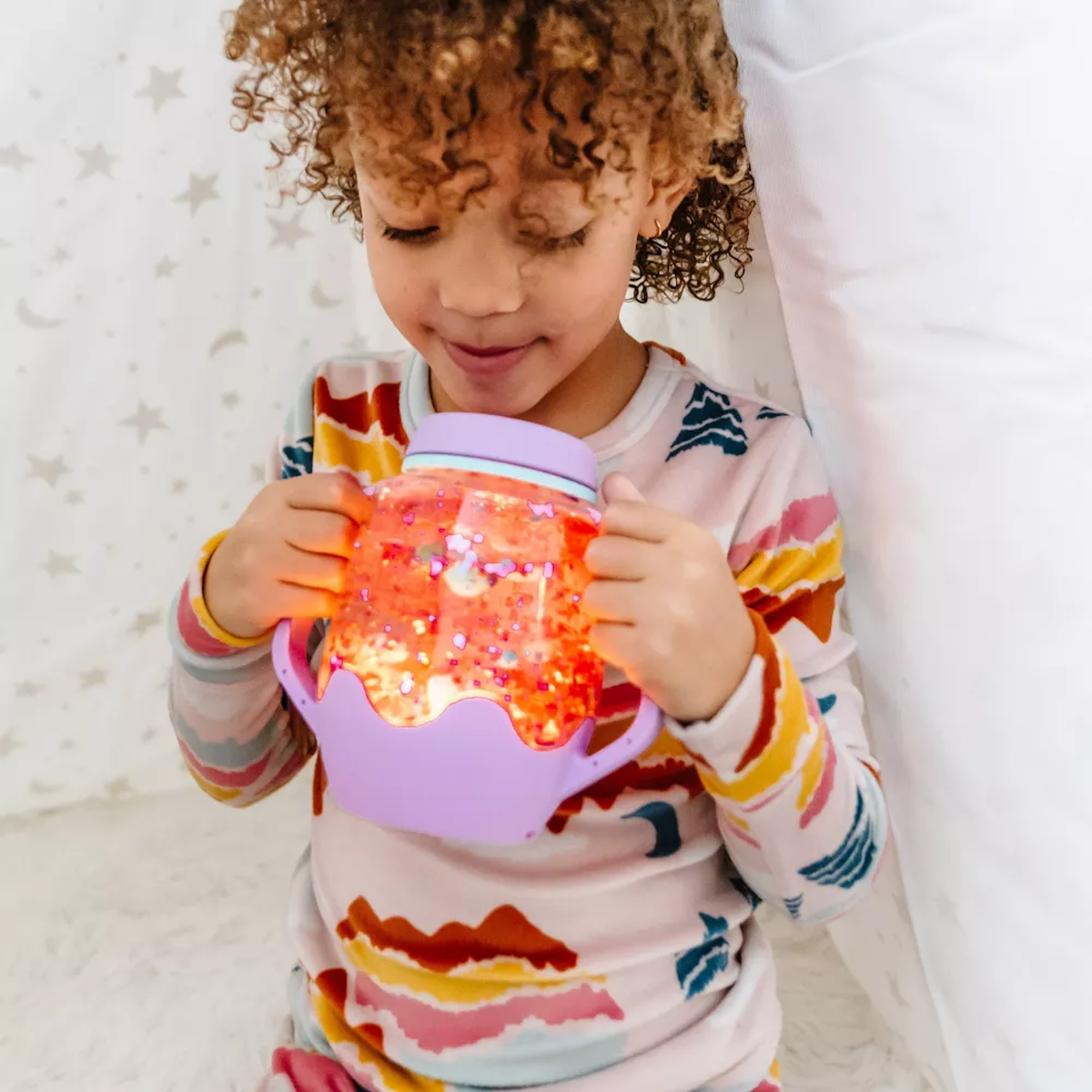 Child holding and looking down at a glowing orange-lit Glo Pals sensory play jar with a purple base.