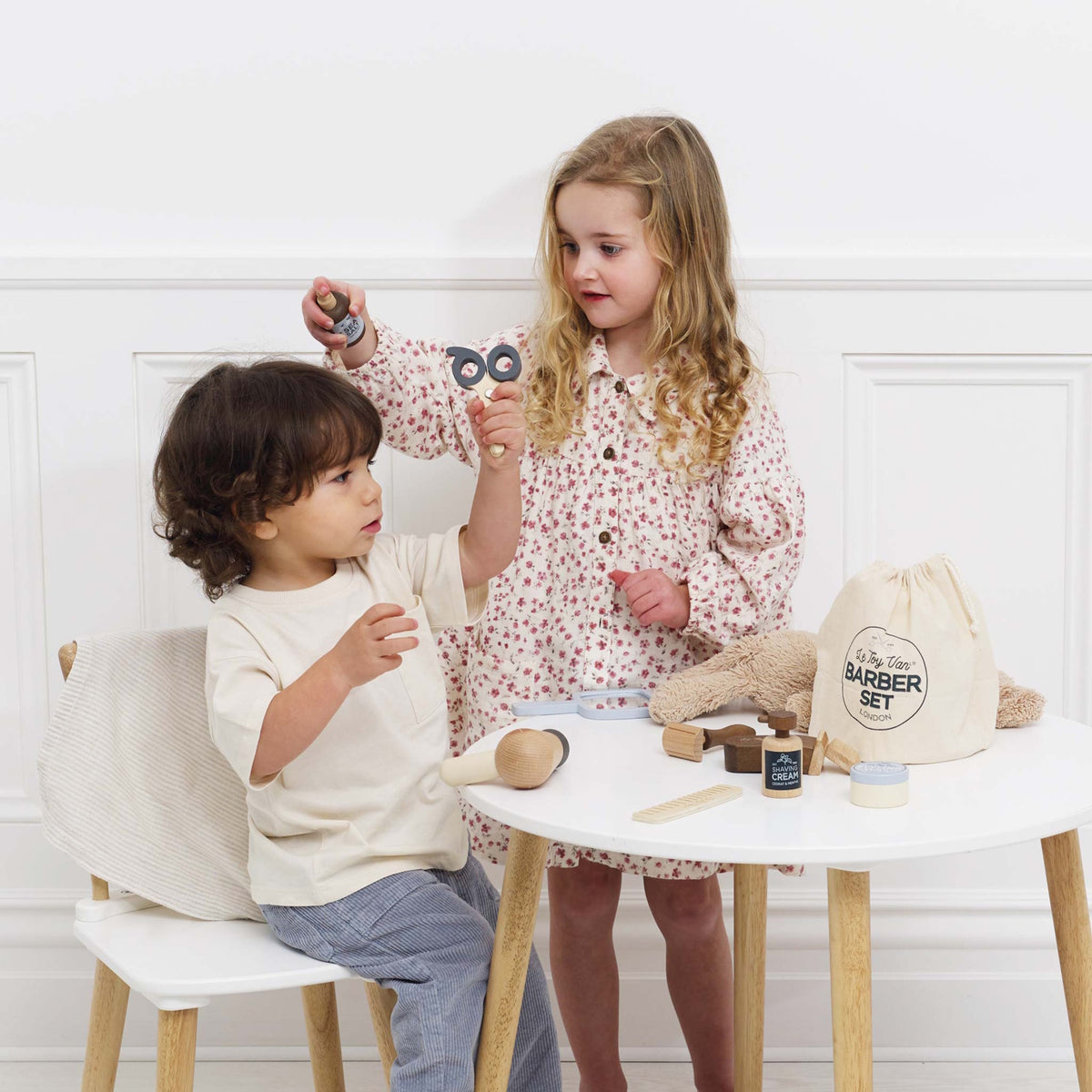 Two young children playing with a wooden hairdresser and barber kit set at a small table.