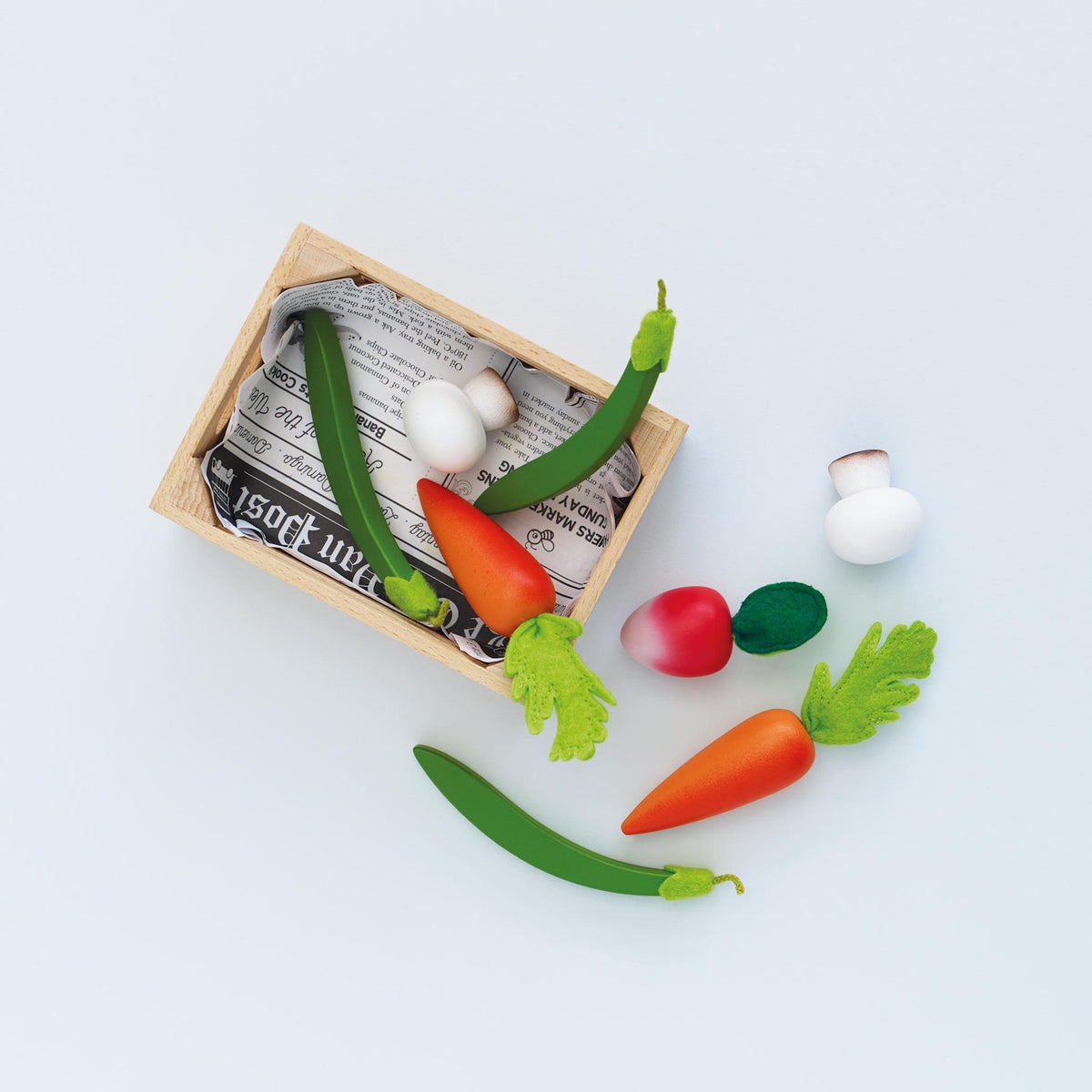 Wooden play vegetables with felt tops arranged beside and inside a newspaper-lined crate.