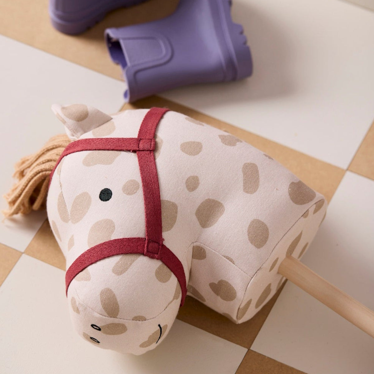 A polka dot hobby horse with a red harness resting on tiled flooring next to purple rain boots.