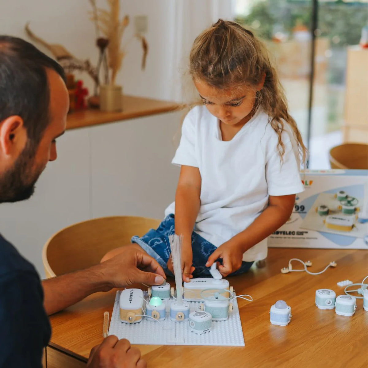 A young girl and a man building an educational electronics kit together at a wooden table.