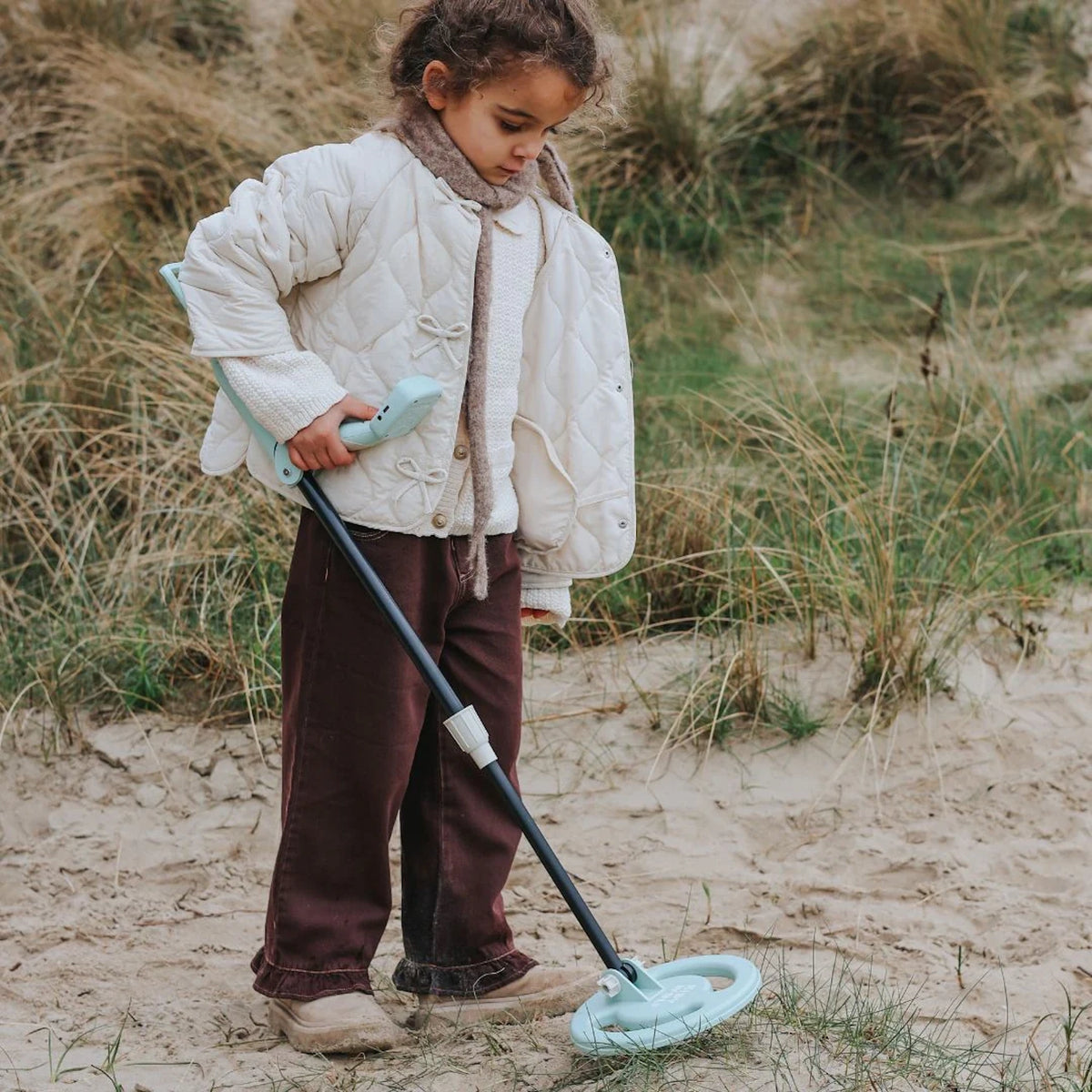 A young child using a light blue toy metal detector on a sandy, grassy beach.