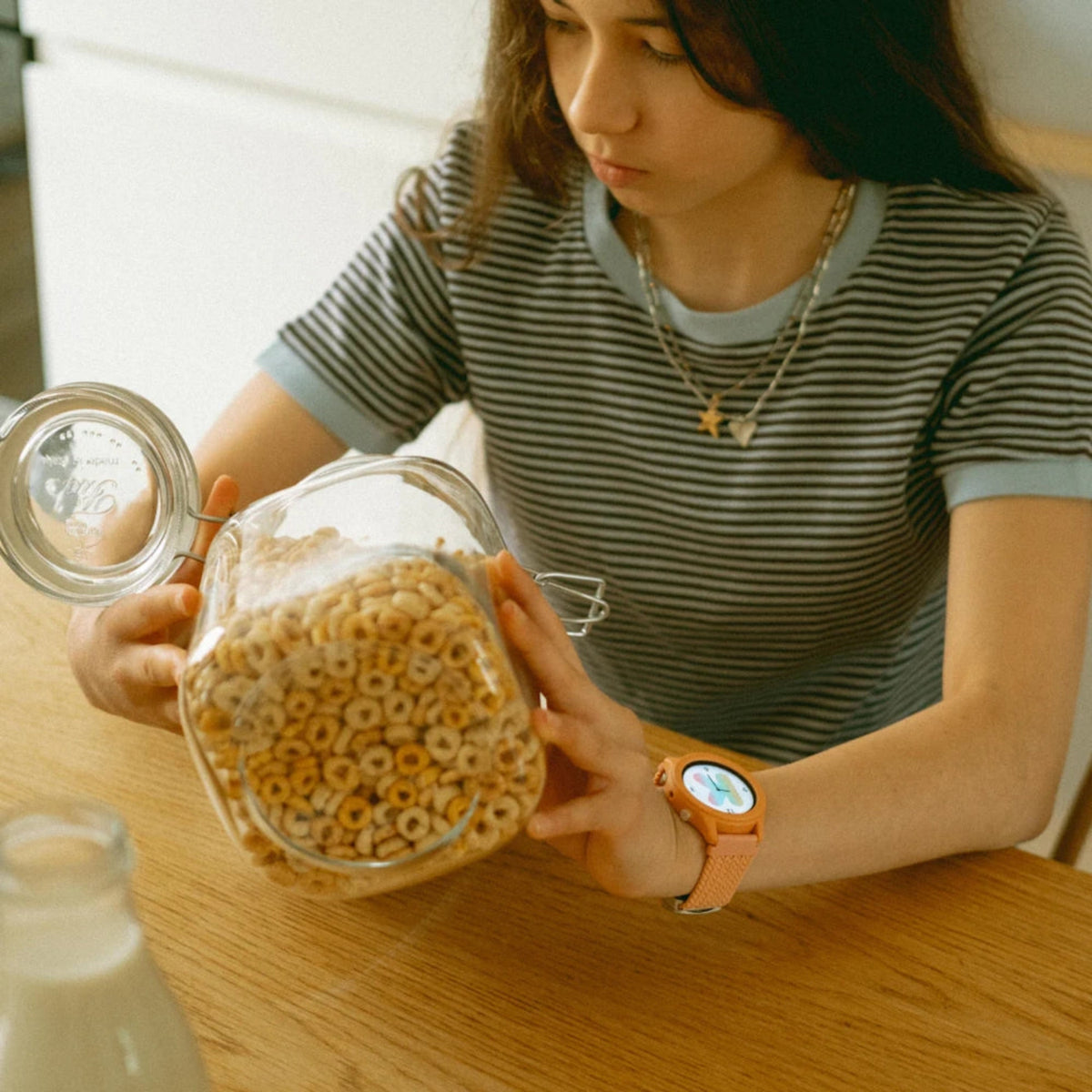 A person wearing an orange smartwatch holds a large glass jar filled with cereal at a wooden table.