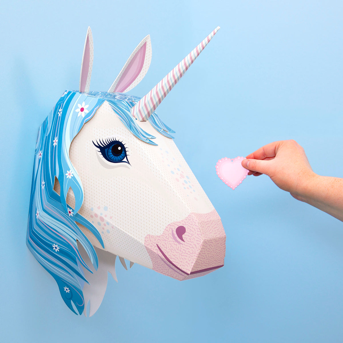A hand holds a pink felt heart towards a paper unicorn head with blue mane and striped horn, mounted on a light blue wall, creating a whimsical feel.