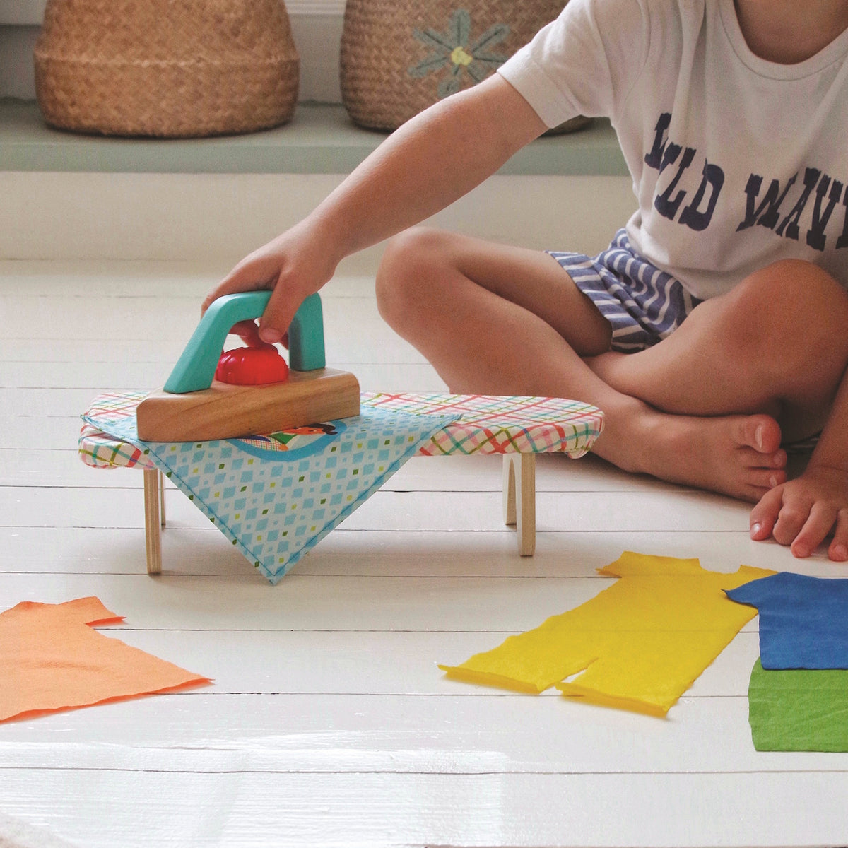 Child using a wooden toy iron and ironing board set on a white floor with colorful fabric clothes.