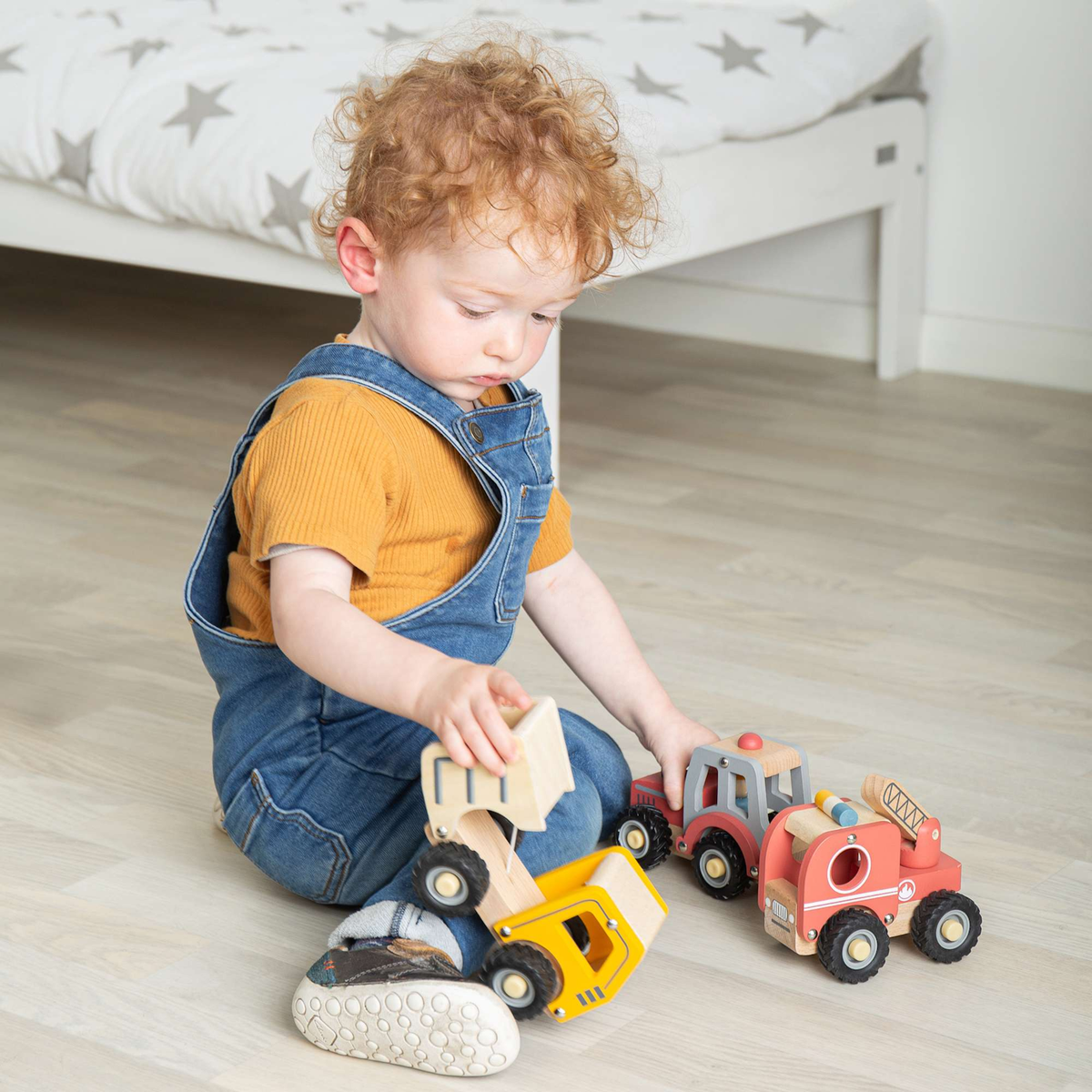A toddler with curly hair, wearing blue overalls and an orange shirt, plays on a wooden floor with colorful toy vehicles, expressing curiosity.