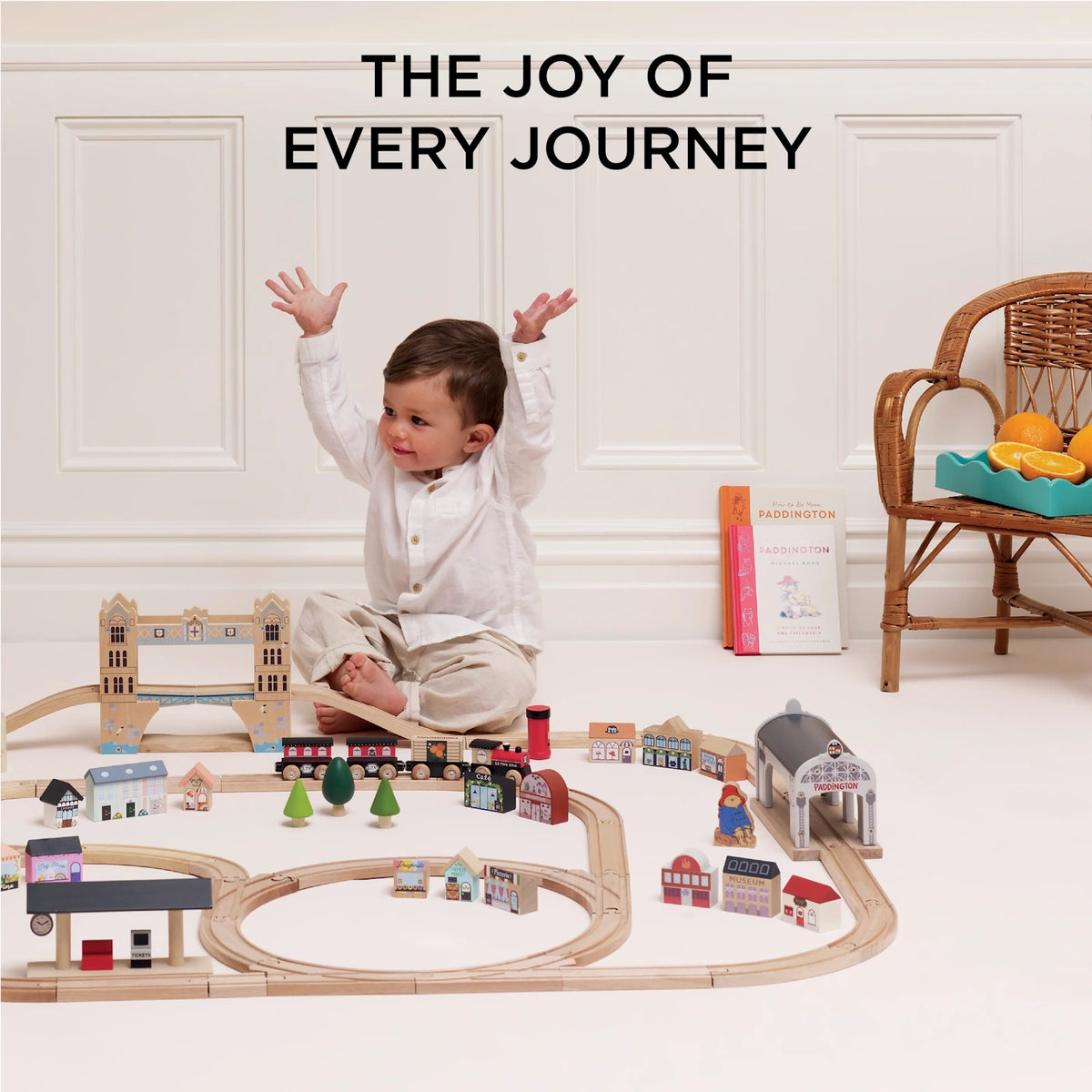 A happy child raising arms while sitting behind an assembled wooden toy train set with Paddington accessories.
