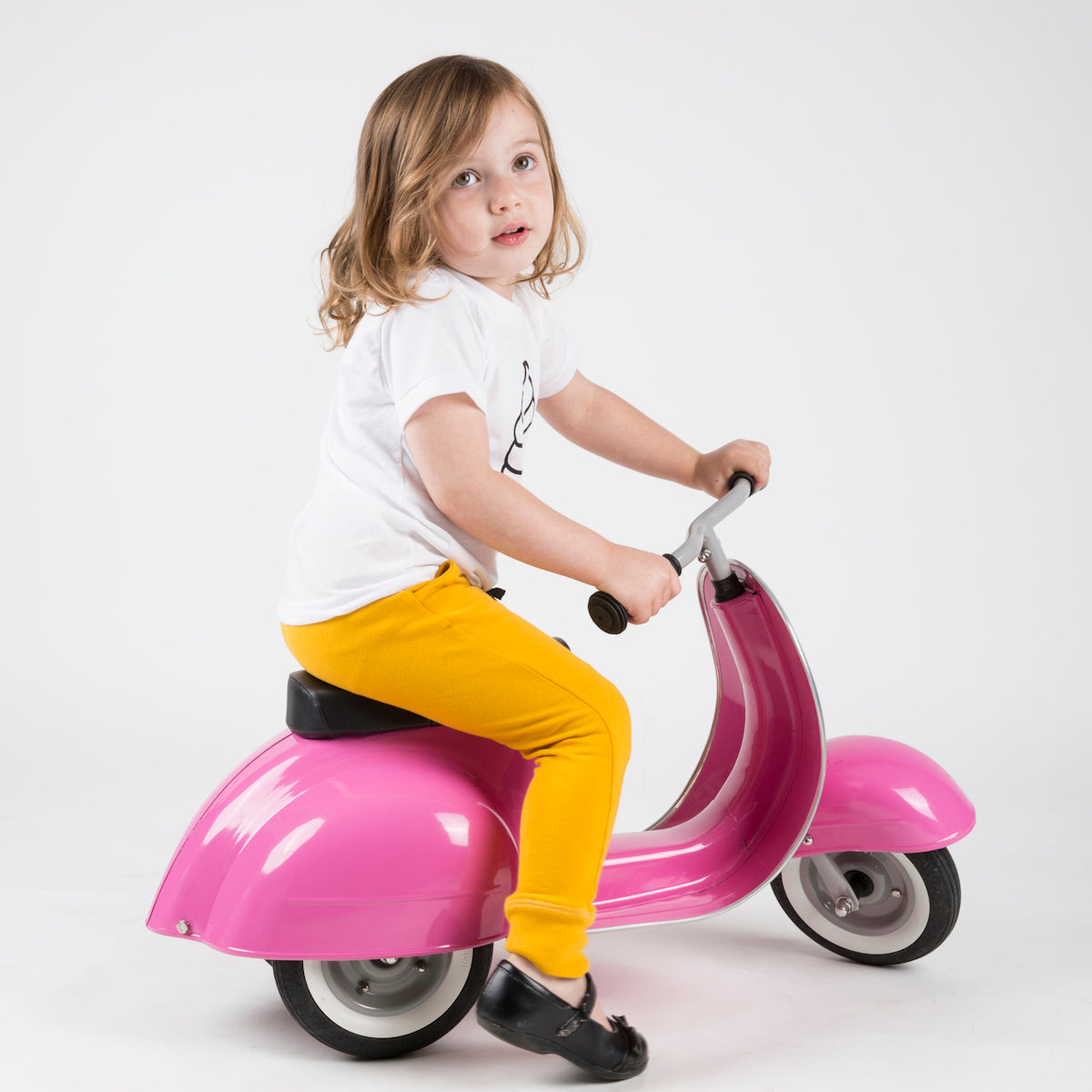 Child in yellow pants and a white shirt rides a small, pink toy scooter indoors. The child looks curious and the scene is playful.