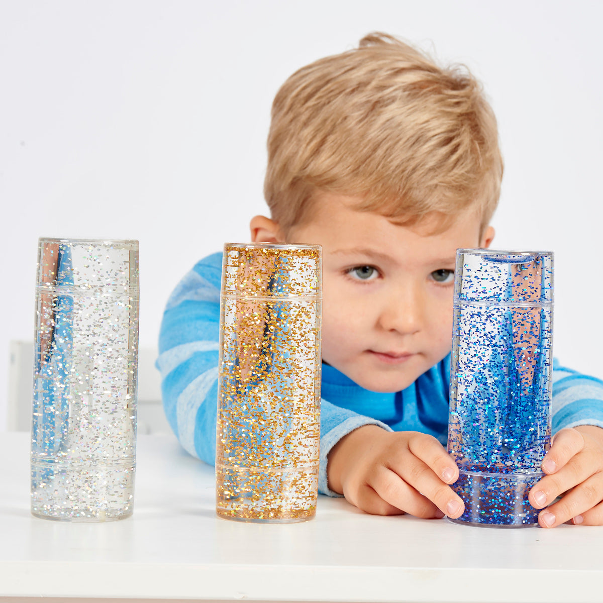 A child in a blue striped shirt is smiling and holding a clear cylinder filled with blue glitter. Two similar glitter cylinders are on a white table.
