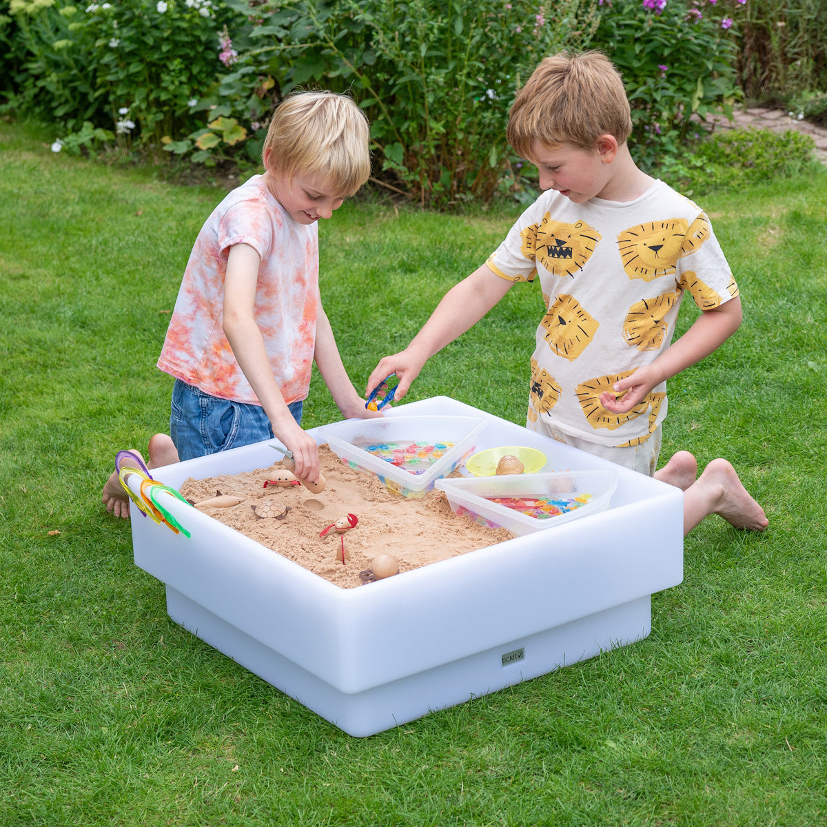 Children plays in a white plastic sandbox on a grassy lawn, stacking colorful cups. Bright toy balls are placed in a container nearby, conveying a playful mood.