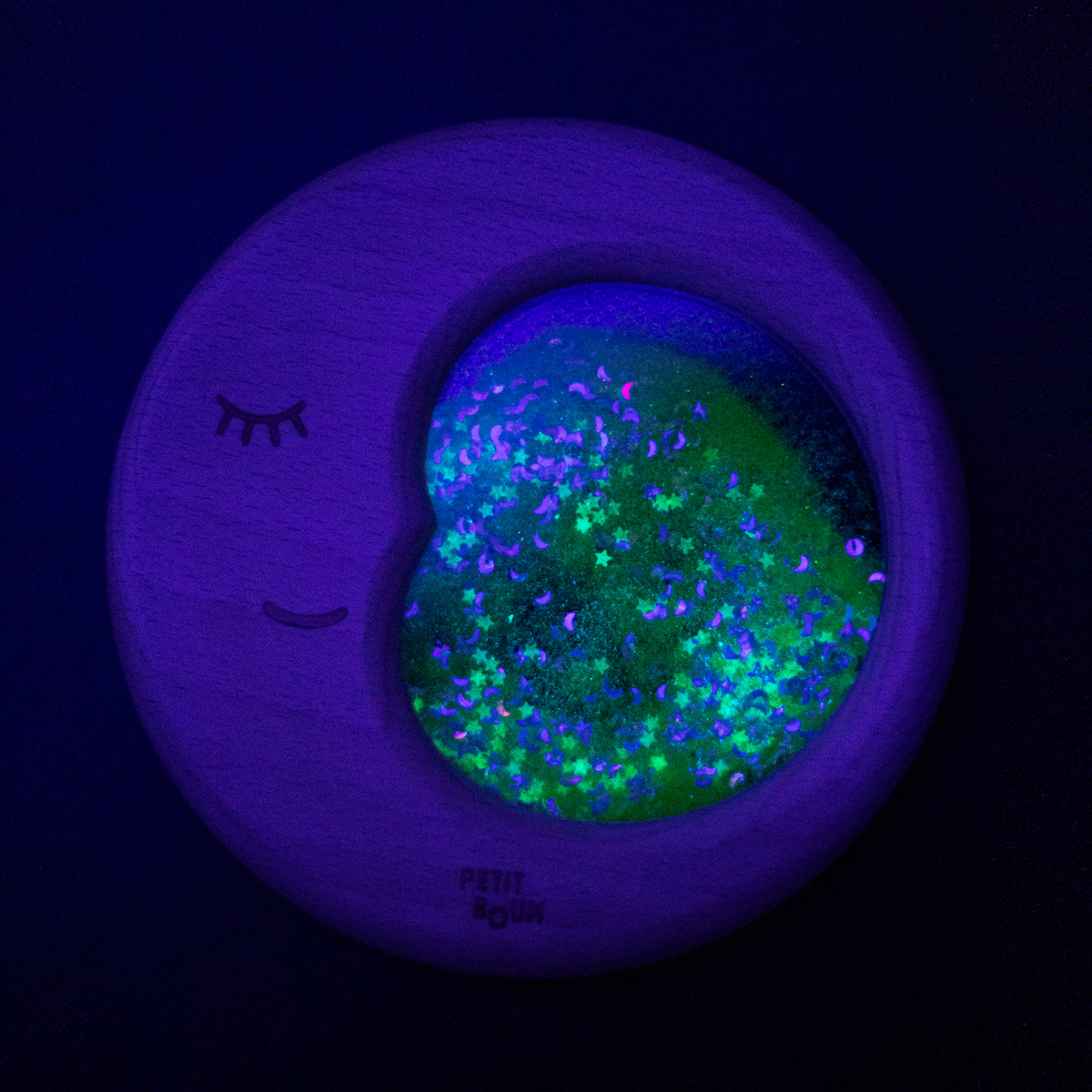 A Petit Boum Flow Moon sensory toy glowing under UV light, showcasing sparkling stars and crescent moons in a dark setting.