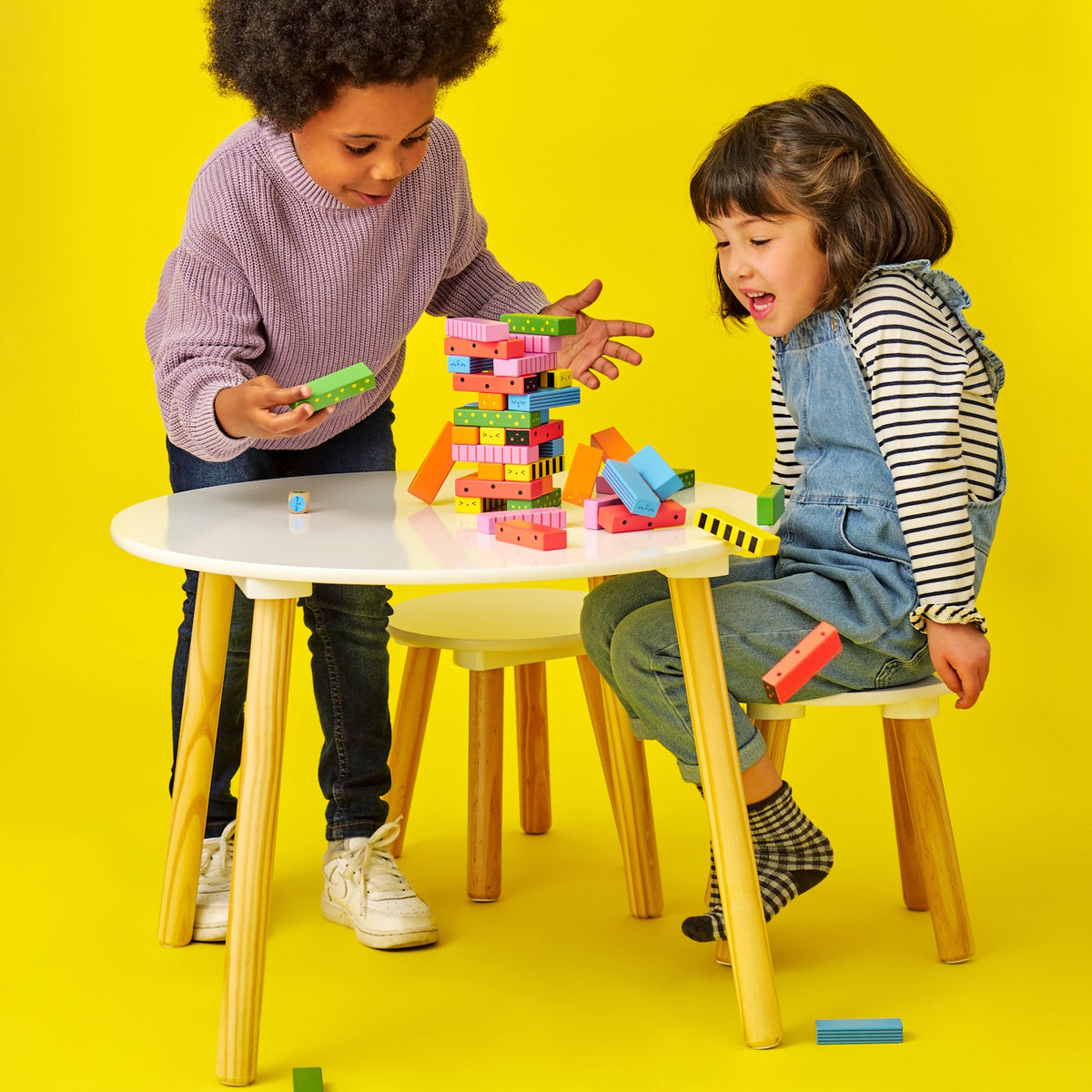 Two children playing a wooden stacking game at a white table, watching colorful blocks fall over against yellow.