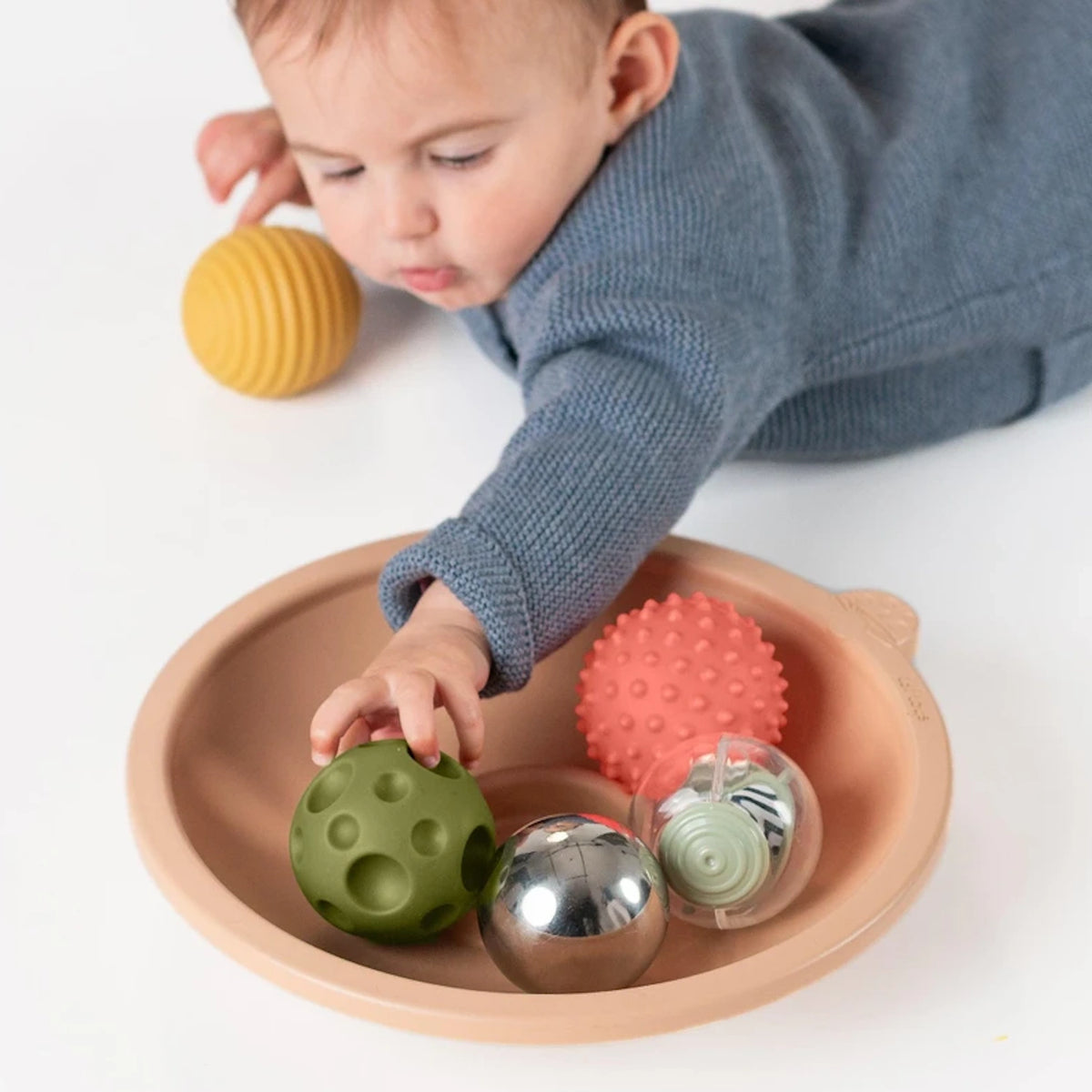 Baby on tummy reaching into a Taf Toys activity tray filled with textured sensory balls.