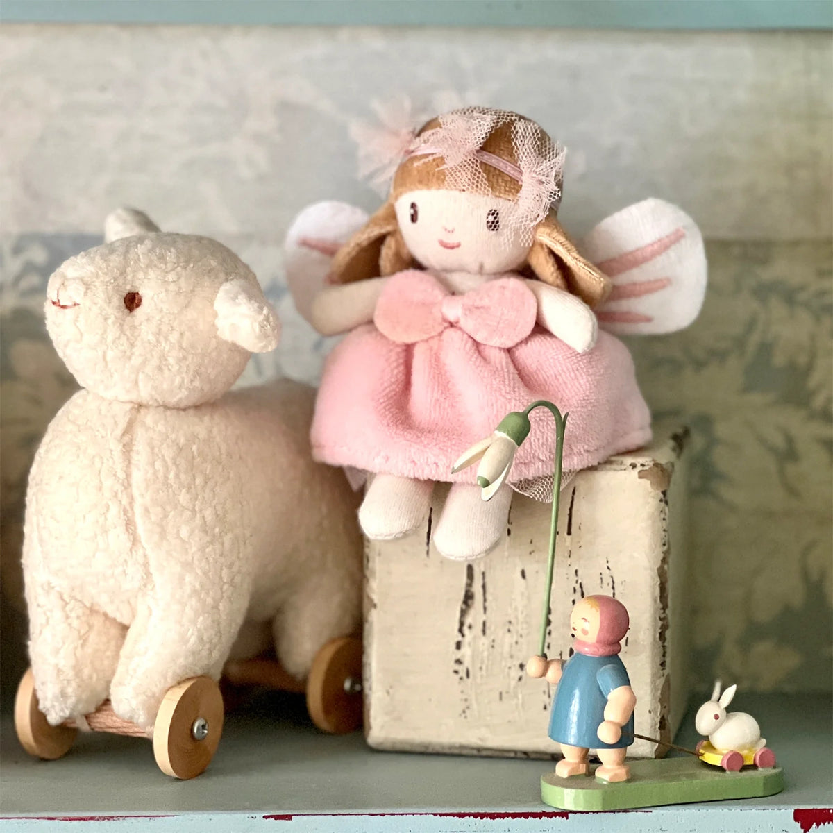 Small plush fairy doll in a pink dress sitting next to a plush toy lamb on wheels.