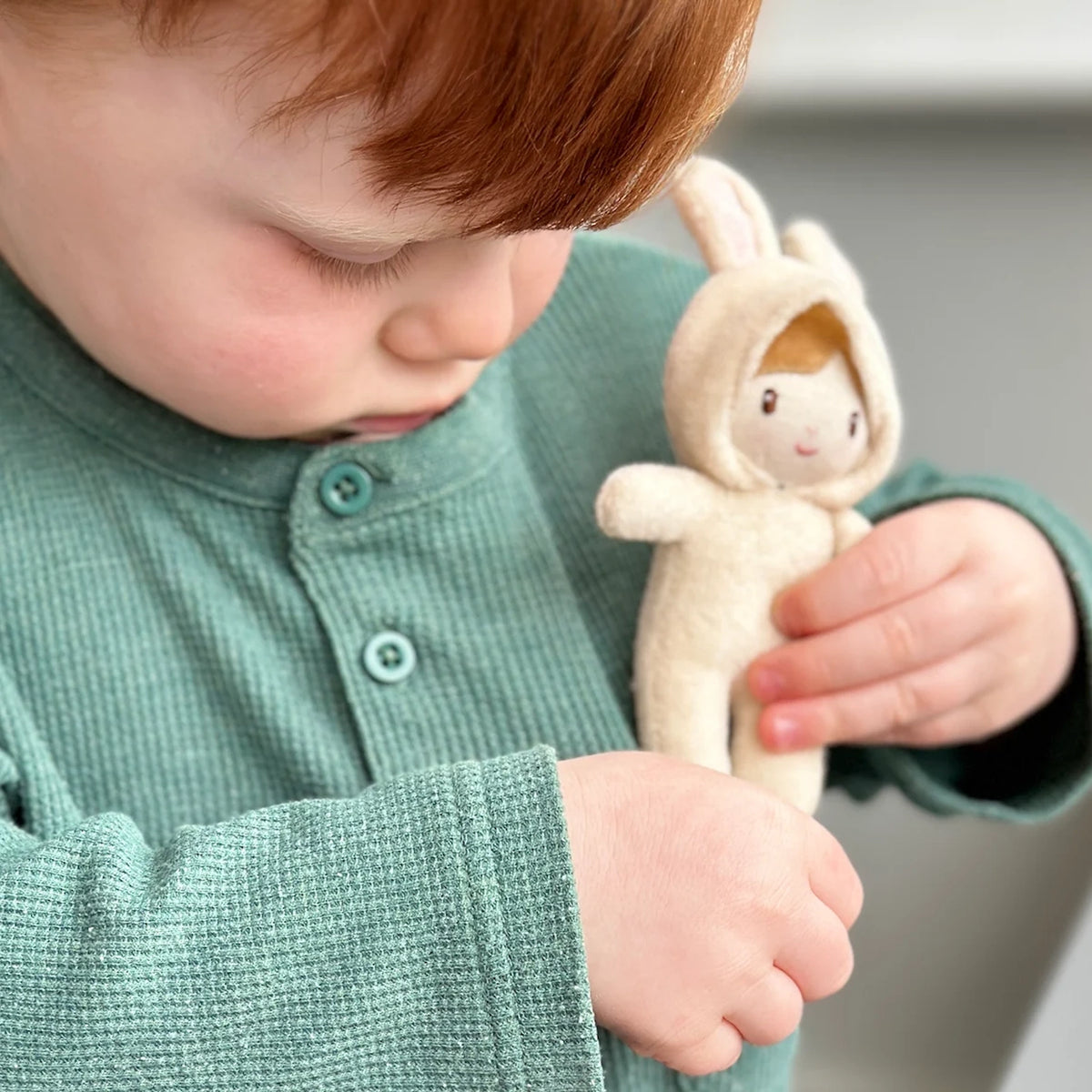 A red-haired toddler wearing a green shirt holding a small cream plush bunny doll close to their chest.