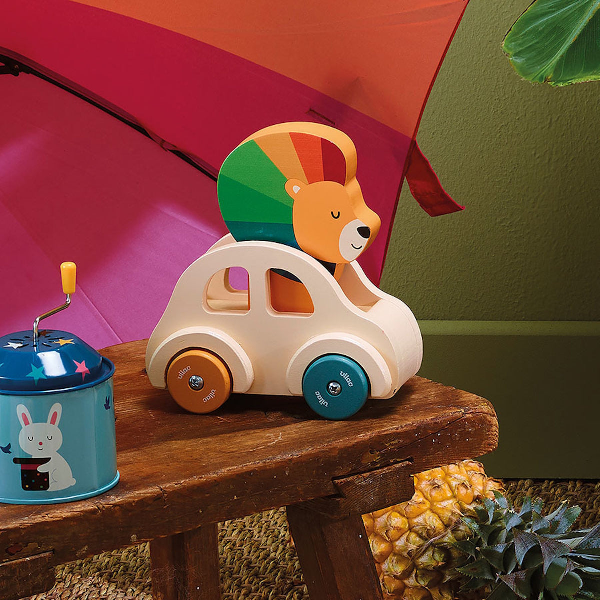 Wooden lion toy car by Vilac on a rustic bench next to a pineapple and a blue rabbit tin.