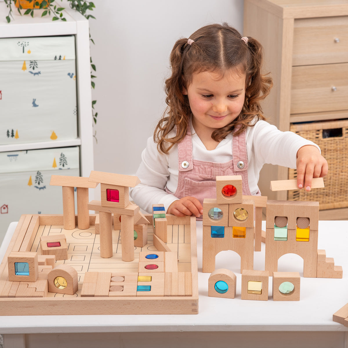 A young child builds a tower with wooden blocks featuring colorful transparent windows. The setting is bright and cheerful, promoting creativity and concentration.