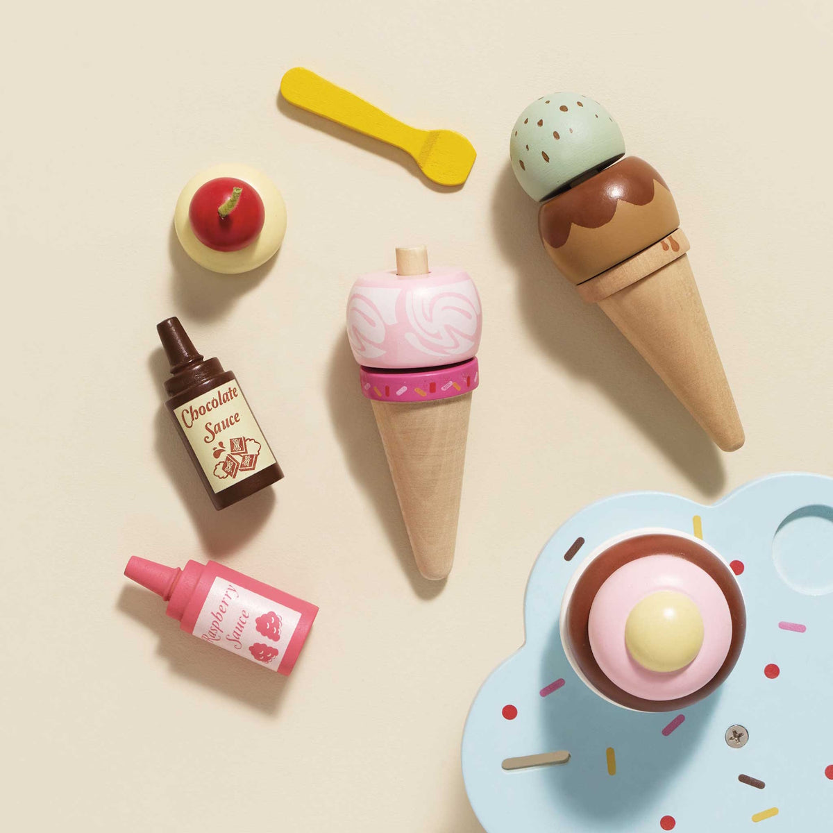 Top-down view of wooden play ice cream cones, scoops, toppings, and sauce bottles on a neutral background.