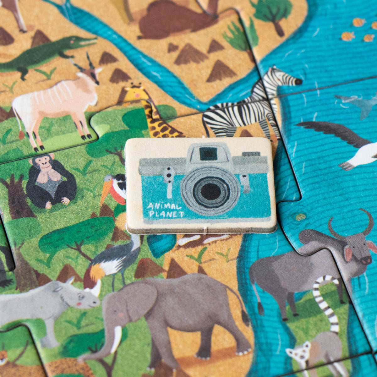 A close-up of an Animal Planet puzzle piece featuring a blue camera token placed on a vibrant illustration of the African savanna, surrounded by elephants, rhinos, and zebras.