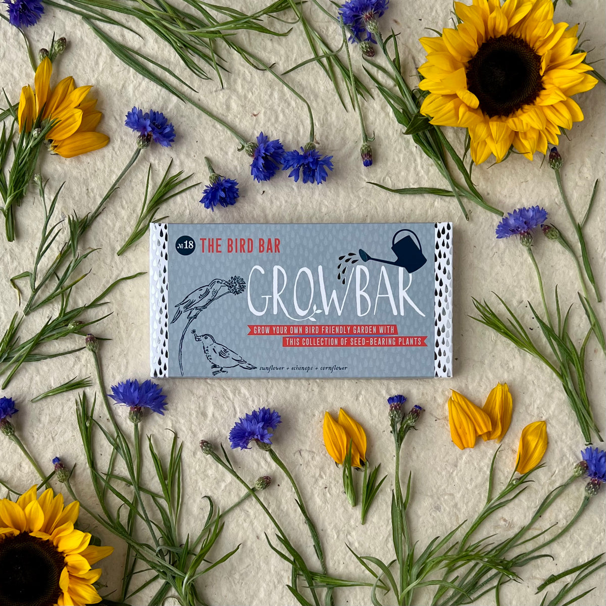 A grey Growbar package labelled "The Bird Bar," surrounded by bright yellow sunflowers, blue cornflowers, and lush greenery, ideal for growing bird-friendly plants.