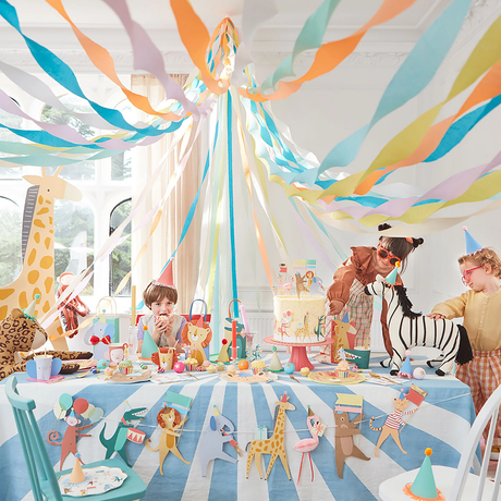 Children sit at a party table decorated with colorful streamers and an animal parade garland.
