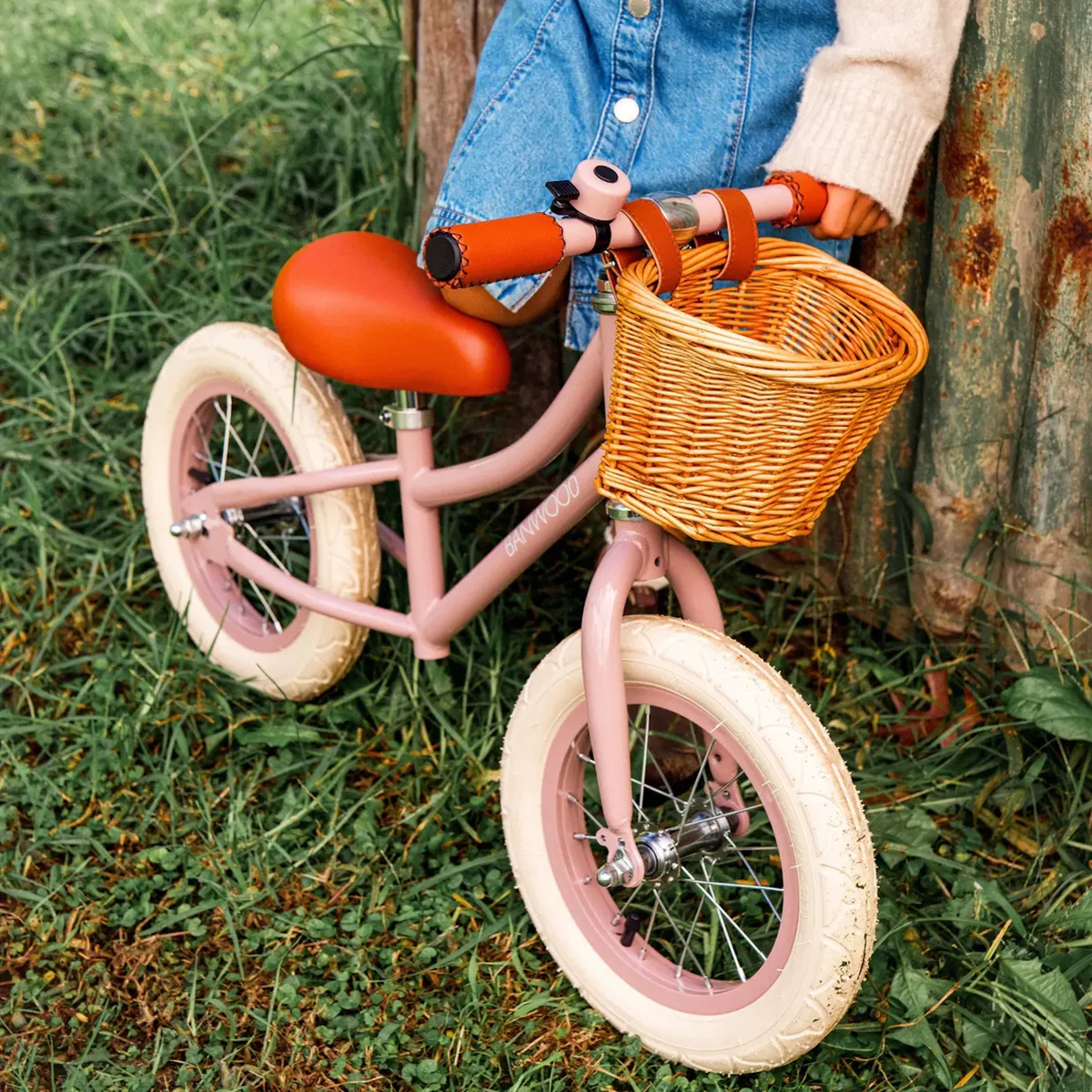 A pink balance bike with a wicker basket and cream tires rests against a rustic wall. A child in a denim skirt and sweater stands nearby on grass.