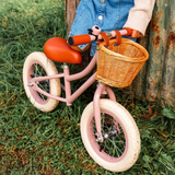A pink balance bike with a wicker basket and cream tires rests against a rustic wall. A child in a denim skirt and sweater stands nearby on grass.