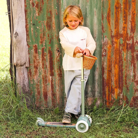 Young child standing with a sage green 3-wheeled scooter against a rustic, rusted metal wall outdoors.