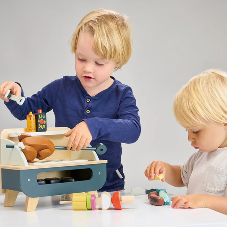 Two children playing with a wooden toy barbecue grill and play food.