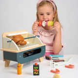 Young girl holding a wooden play food skewer next to a toy barbecue set.