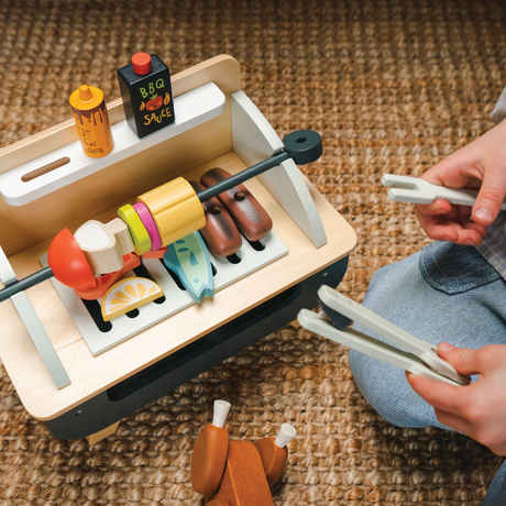 Overhead view of a child using wooden tongs to play with a barbecue set on a woven rug.