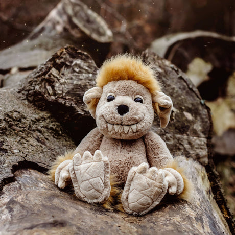 A smiling beige plush creature toy with fluffy yellow hair sitting outdoors on a large tree log.