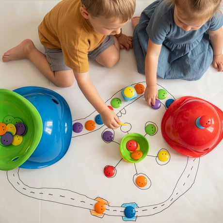 Two children playing with Bilibo Game Box toys on a hand-drawn board, featuring colorful mini shells, dots, and a pixel die.
