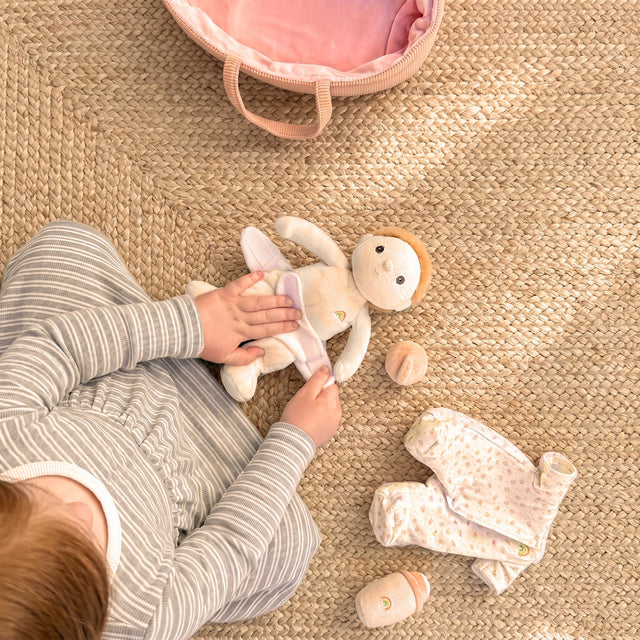 A child's hands changing a Dinky Dinkum doll on a woven rug next to doll clothes and a pink bag.