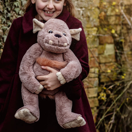 A smiling young girl in a dark red coat holding a soft, plush grey monster toy outdoors.