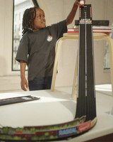A child plays with a Candylab Roadworks Hairpin Turn kit, racing a toy car down a steep wooden track ramp from a stool.